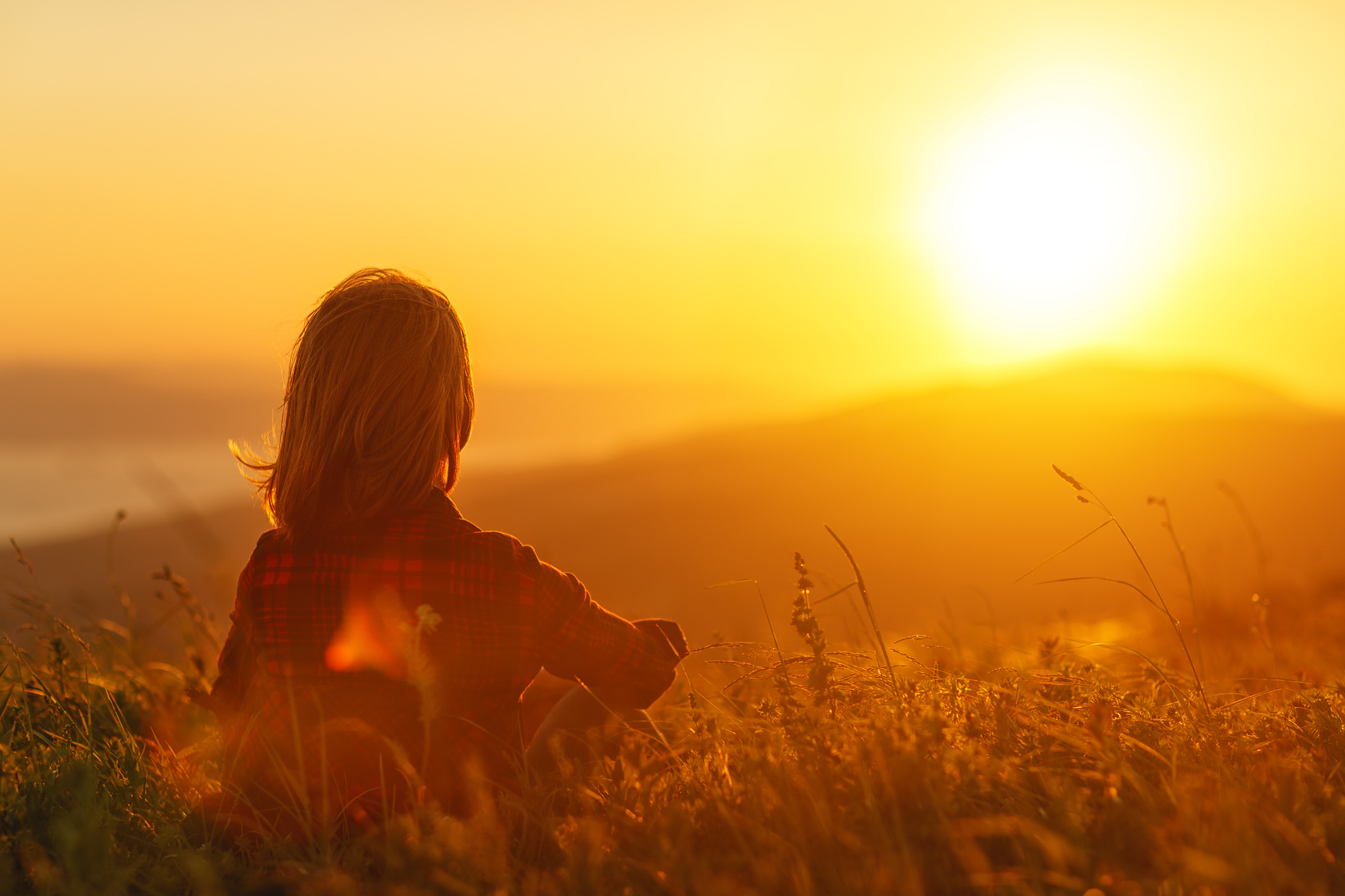 Woman sits with her back in the field and look sunset in the mountains