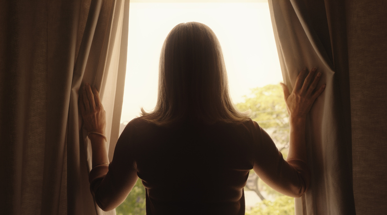 A mature woman opening the curtains in her bedroom during a relaxing morning at home. Rear view of a senior lady in silhouette looking out the window, starting the day