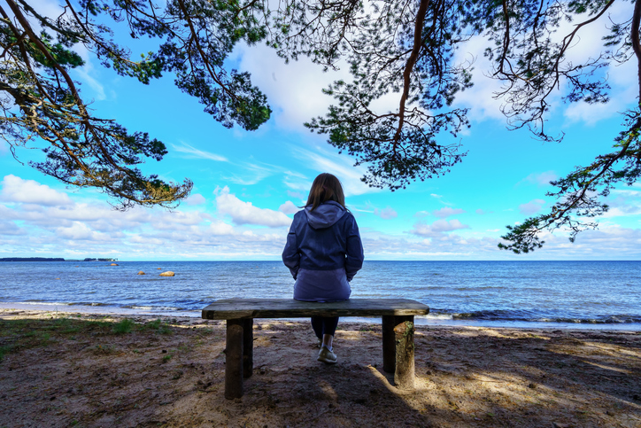 Woman from behind sitting on a bench facing the sea and tree branches around her.