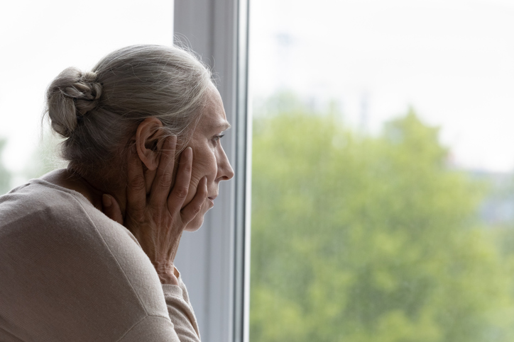 Upset lonely senior lady looking out of window with despair