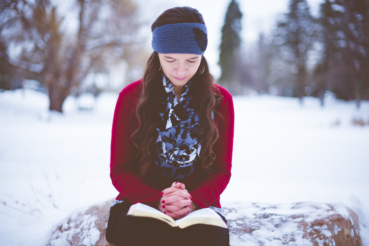 Beautiful female with a purple jacket reading the bible with her hands clenched sitting on a stone