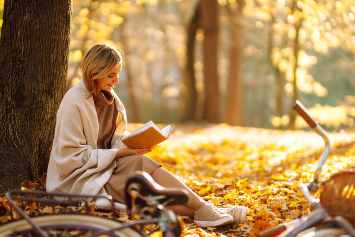 Beautiful young woman sitting on a fallen autumn leaves in a park, reading a book.