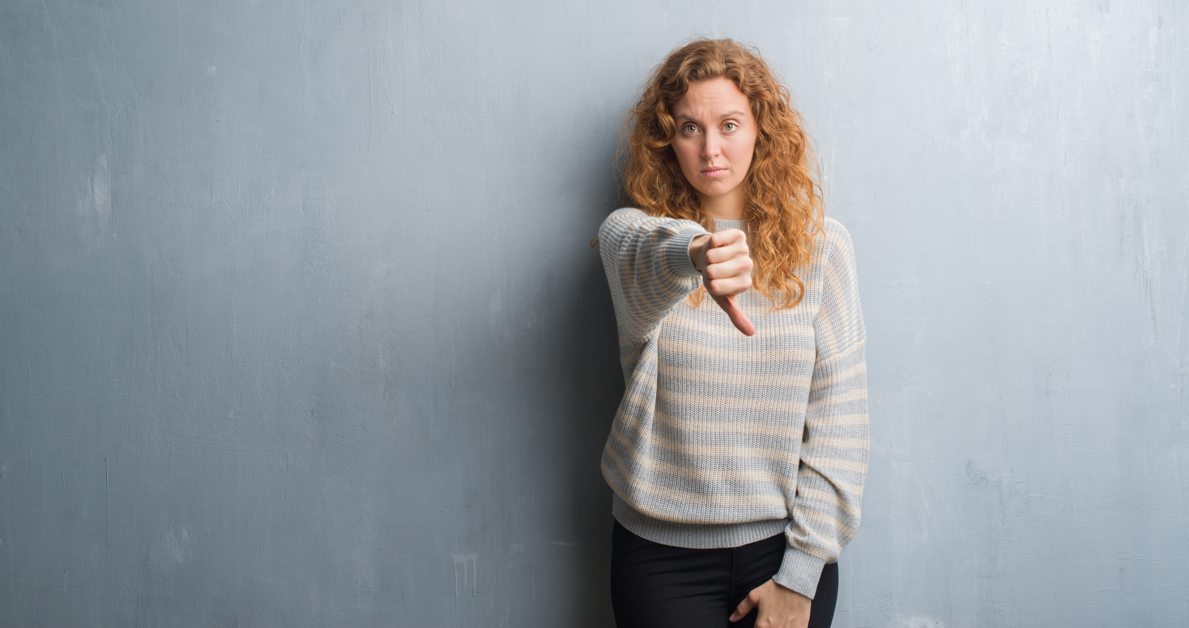 Young redhead woman over grey grunge wall looking unhappy and angry showing rejection and negative with thumbs down gesture. Bad expression.