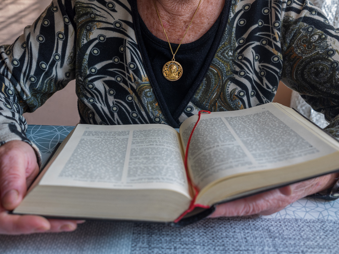 Older woman reading the bible with a gold medallion of the Virgin Mary hanging around her neck.