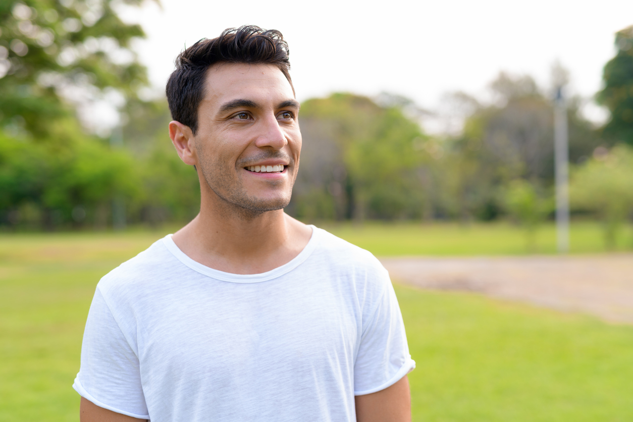 Face of happy young handsome Hispanic man thinking at the park