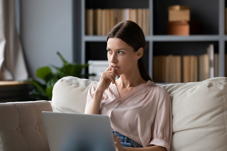Thoughtful woman holding laptop, pondering ideas, sitting on couch at home