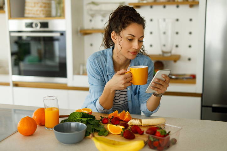 Young woman using her mobile phone while preparing fruit smoothie in the kitchen
