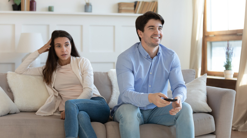 Smiling man watching tv, bored angry woman sitting separately