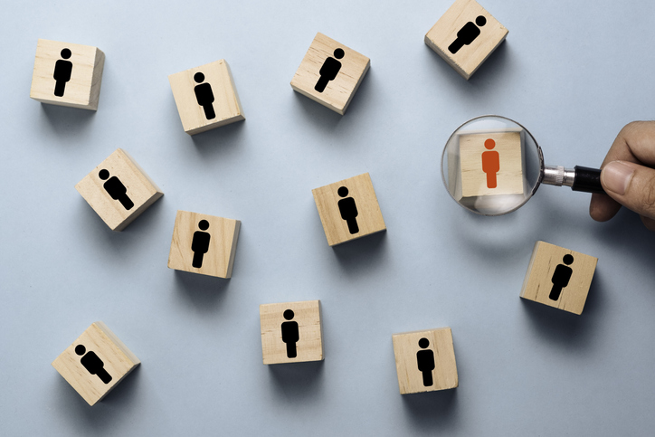 Hand holding magnifier glass to searching red human icon on wooden cube block among black people icon. Human development concept.