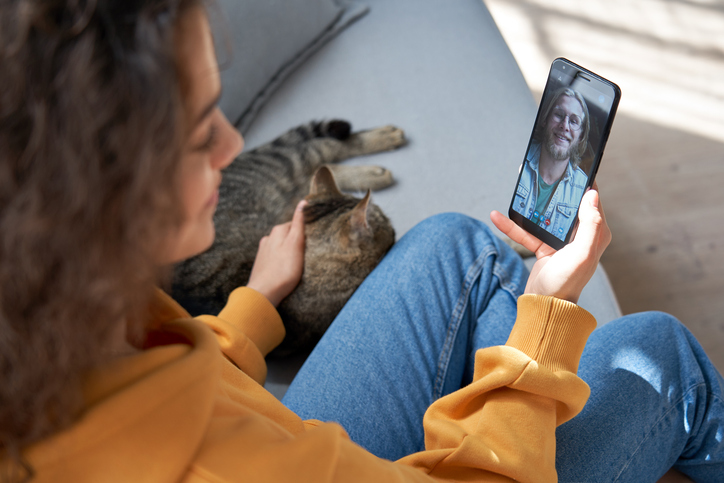 Young hispanic latin teen girl sit on sofa with cat at home holding phone video calling distance friend dating online on mobile screen using smartphone videochat zoom app. Over shoulder closeup view