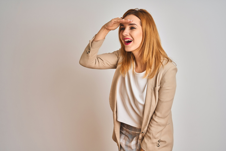 Redhead caucasian business woman standing over isolated background very happy and smiling looking far away with hand over head. Searching concept.