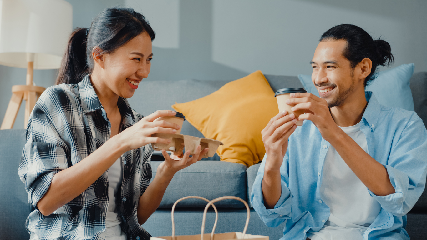 Happy asian young attractive couple man and woman sit at new home drink coffee relax and talk smile with carton package box storage to move in new house.