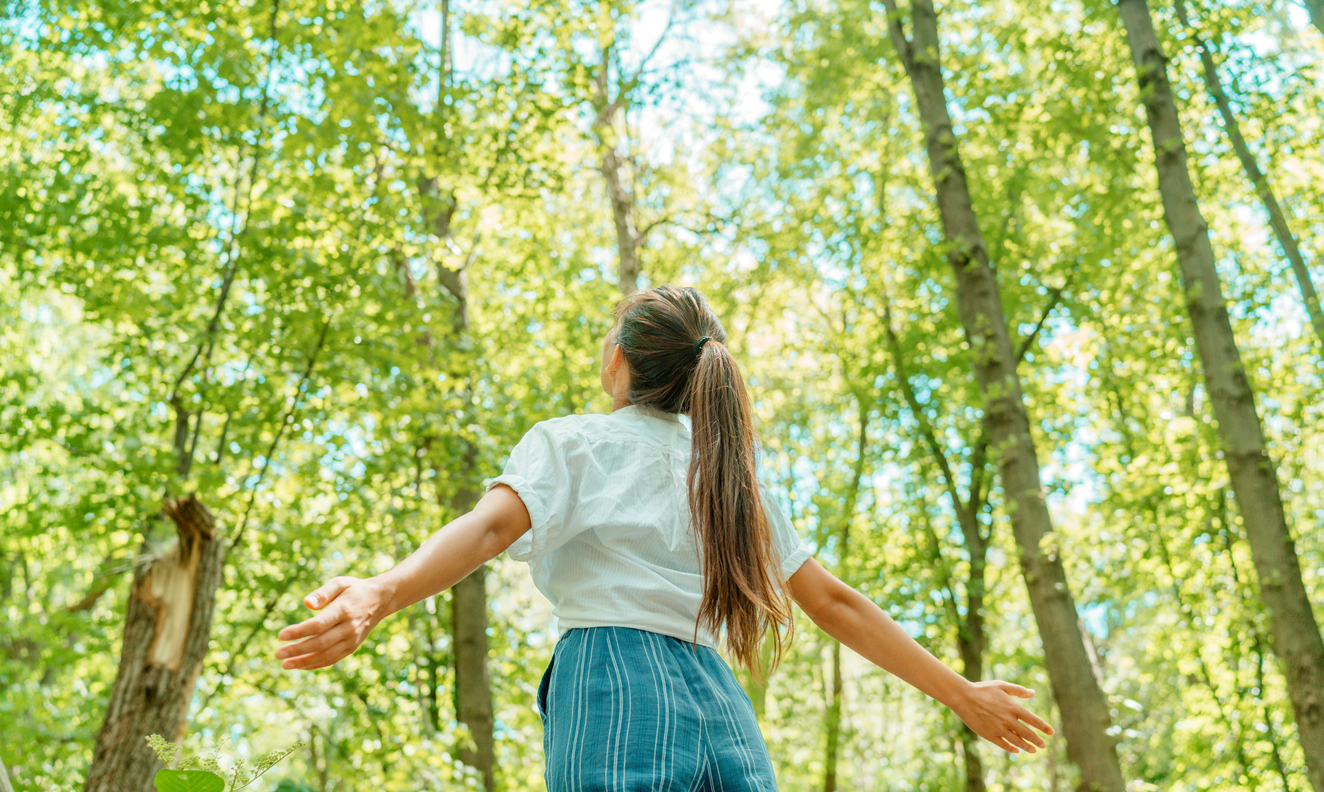 Free woman breathing clean air in nature forest. Happy girl from the back with open arms in happiness. Fresh outdoor woods, wellness healthy lifestyle concept.