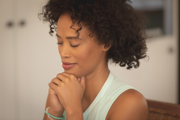 African American woman with hand clasped and eyes closed praying at dining table