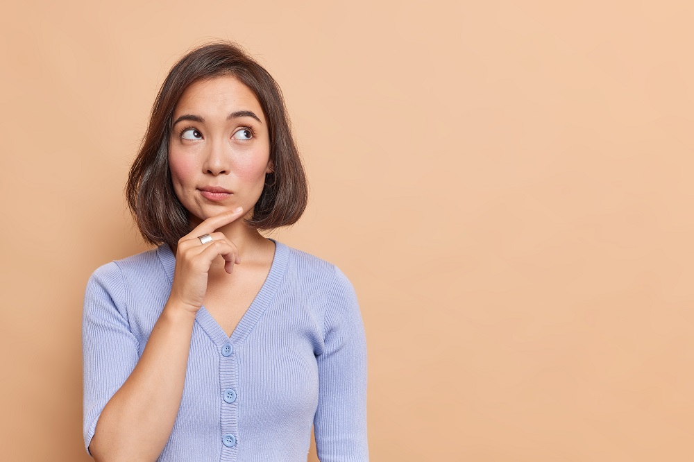 Thoughtful,Asian,Woman,Keeps,Hand,On,Chin,Looks,Pensively,Above