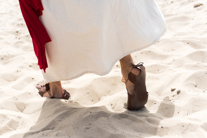 cropped image of Jesus in robe, sandals and red sash walking on sand in desert