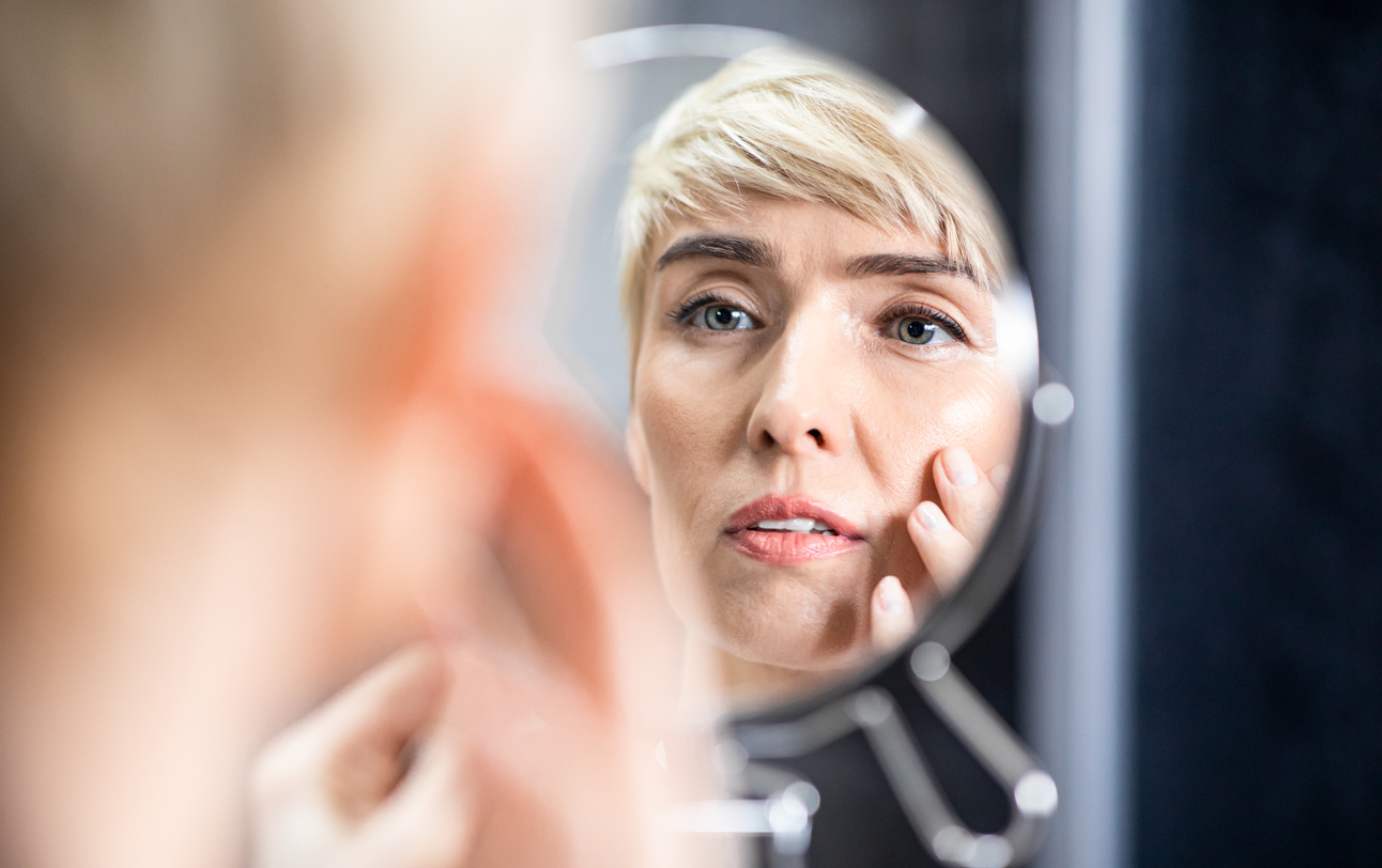 Mature Woman Looking In Mirror Touching Face Standing In Bathroom