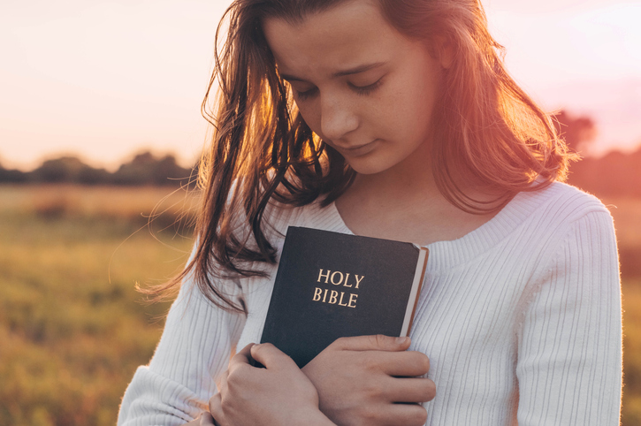 Christian teenage girl holds bible in her hands. Reading the Holy Bible in a field during beautiful sunset. Concept for faith, spirituality and religion