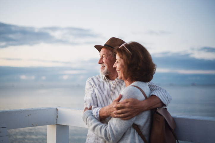 Portrait of happy senior couple in love hugging outdoors on pier by sea, looking at view, summer holiday.