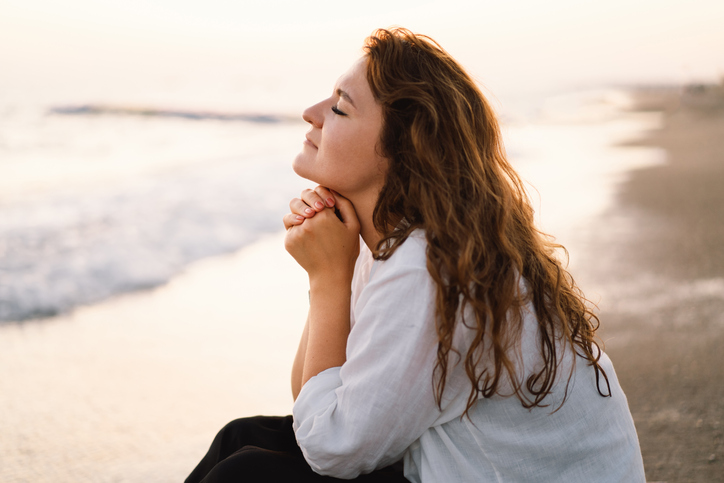 Woman closed her eyes, praying on a sea during beautiful sunset. Hands folded in prayer concept for faith, spirituality and religion.