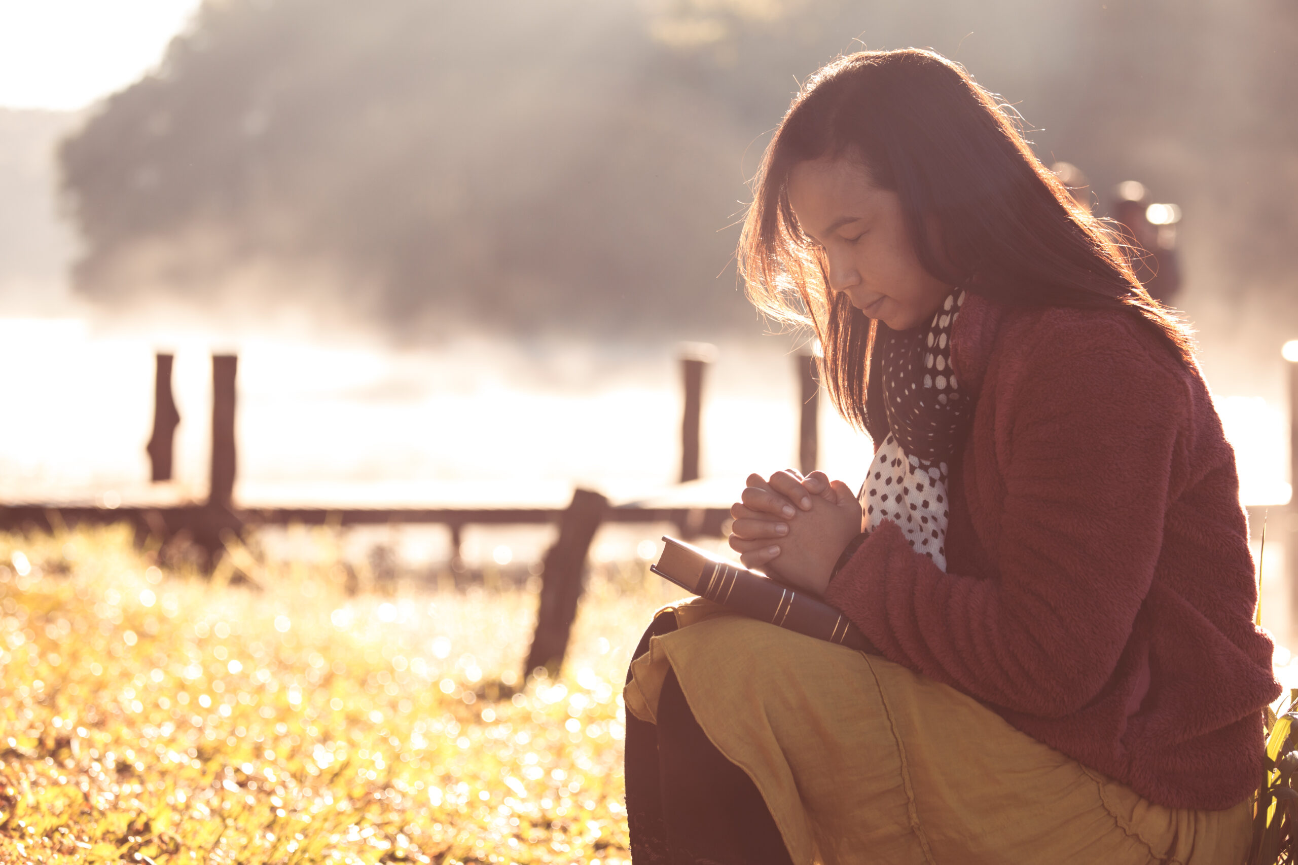 Woman hands folded in prayer on a Holy Bible for faith in beautiful nature background with sunlight