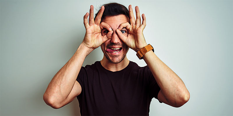 Young handsome man with tattoo wearing purple casual t-shirt over isolated white background doing ok gesture like binoculars sticking tongue out, eyes looking through fingers. Crazy expression.