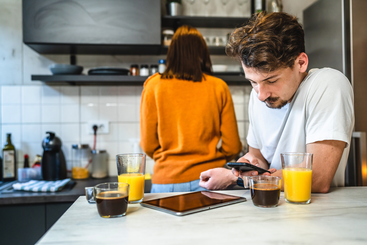 Man use mobile phone in the kitchen while his girlfriend is making breakfast &#8211; husband and wife young couple daily routine concept