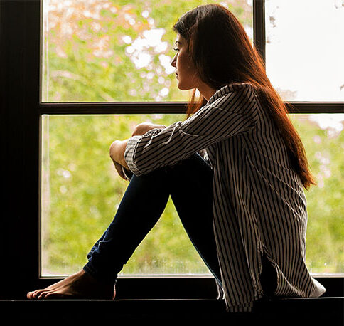 Thoughtful girl sitting on sill embracing knees looking at window