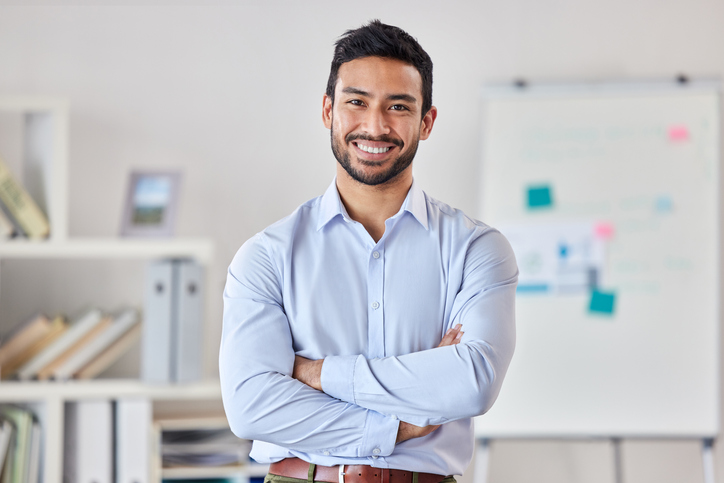 Young happy mixed race businessman standing with his arms crossed working alone in an office at work. One expert proud hispanic male boss smiling while standing in an office