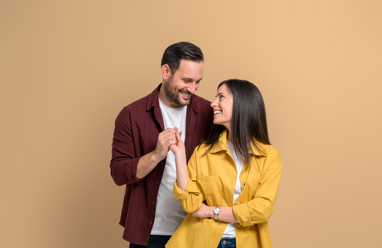 Romantic young man and woman holding hands and smiling while standing over beige background. Attractive couple dressed in casuals looking at each other affectionately