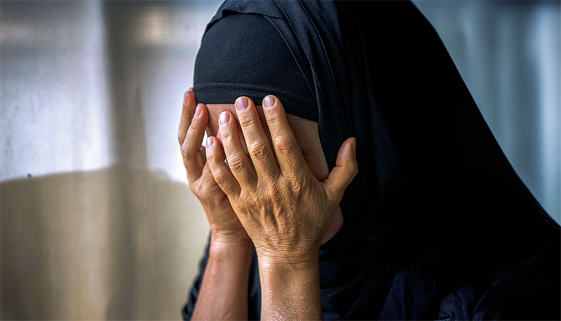 Sad muslim woman looking through a window covering her face with her hands