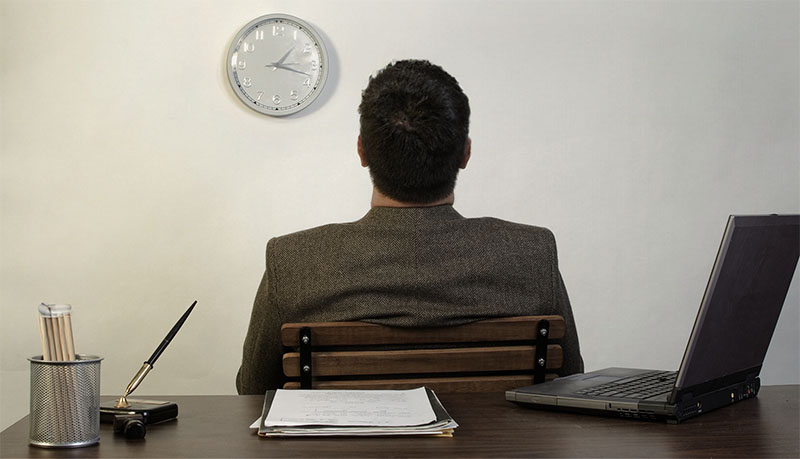 A man sitting backing against a desk looking up at a clock