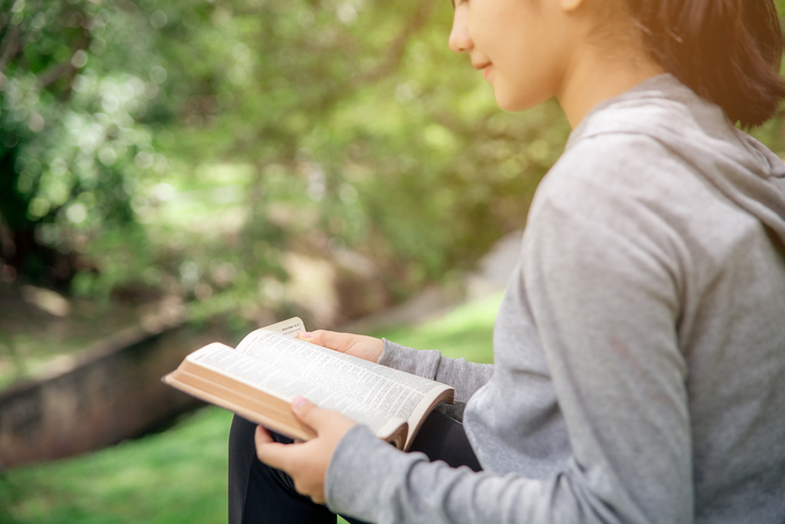 A young Asian woman with a ponytail hairstyle is happily reading a book in the park.