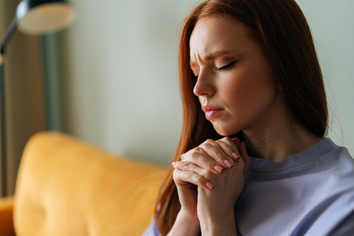 Closeup face of worried anxious young red-haired woman sitting on couch praying god with folded hands having life crisis. Nervous female trying to concentrate