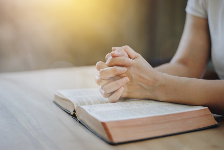 Close up of a woman hands praying on an open bible on a wooden table with window light Bokeh.