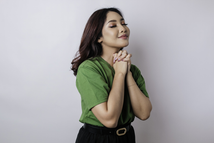 A portrait of Asian woman wearing green t-shirt is praying for success isolated by white background