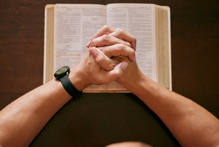 Shot of a young man sitting at a table with his hands clasped in prayer above a bible