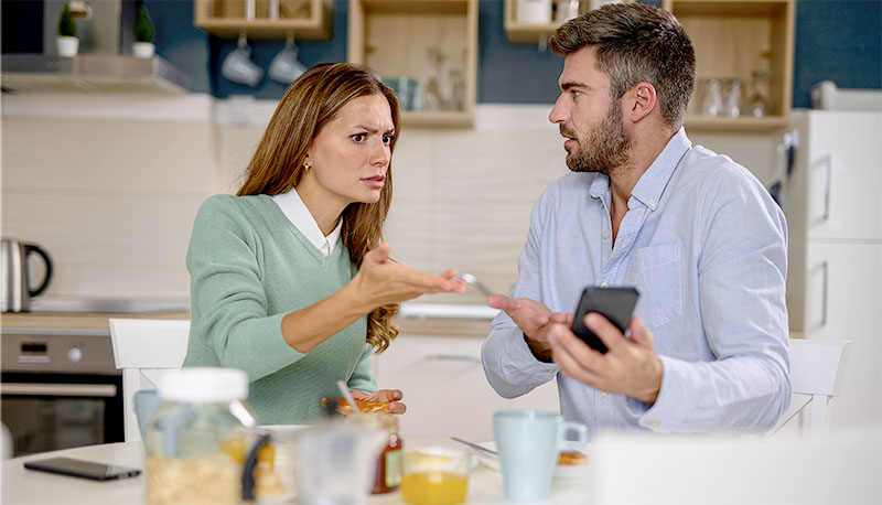Young woman looking irritated while her husband uses his phone during breakfast at home
