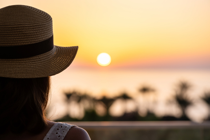 Silhouette of woman in straw hat standing on balcony and looking at sunrise palm sea beach. Rear view