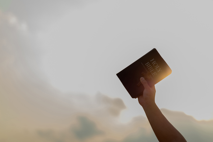 Arm raised into the air with a hand reaching up holding the Holy Bible book and cross on the air. Symbolic posture of prayer and worship.belief, faith.Religious belief concept.