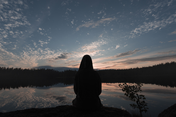 A girl sits on the shore of a forest lake in the evening and admires the beautiful view. View from the rear