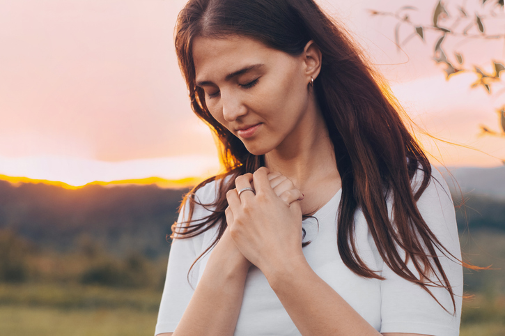 Portrait of a single woman praying and looking down at sunset. Hands folded in prayer concept for faith, spirituality and religion.