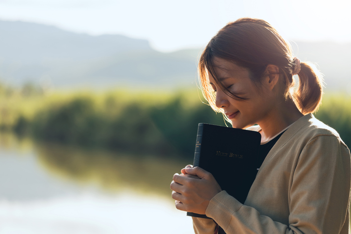 Christian woman praying on the holy bible in a field during a beautiful sunset, holding the bible in her hands. The concept of faith, spirituality, and religion. Peace, hope. Wishing for a better life