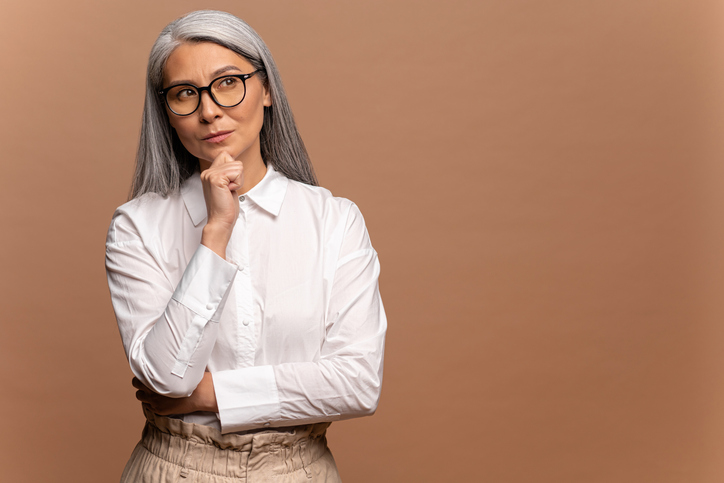 Smart and thoughtful mature woman holding her chin and pondering idea, making difficult decision, looking uncertain doubtful. Indoor studio shot isolated on beige