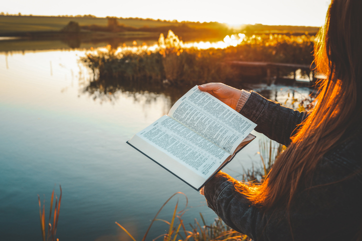 Woman reading open Bible by a peaceful lake during golden sunset, with warm light reflecting on water and long hair glowing in sunlight. Tranquil outdoor moment of faith and reflection