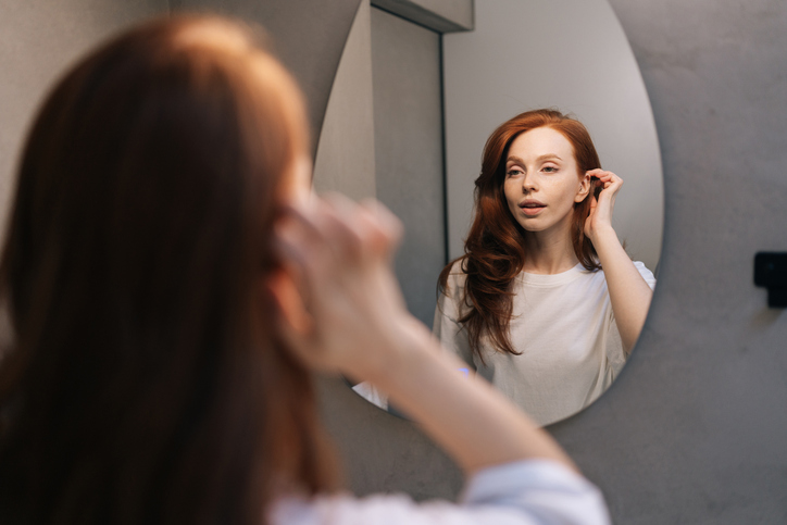 Reflection on mirror of pretty young woman looking in mirror, preens, admires, straightening clothes and hairstyle. Redhead female enjoy beauty routine