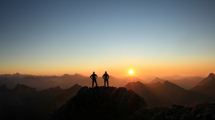 Two Men reaching summit enjoying freedom and looking towards mountains sunset.