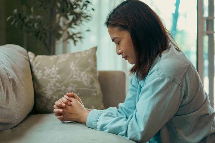 ​Woman sits on a knee and prays in front of a sofa.