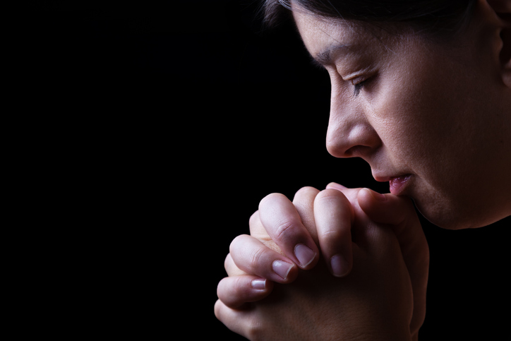 Faithful woman praying, hands folded in worship to god with head down and eyes closed in religious fervor, on a black background.