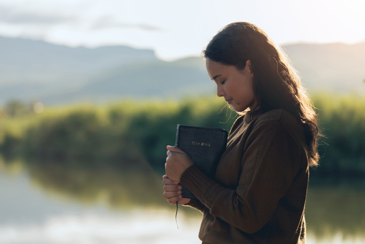Christian woman praying on the holy bible in a field during a beautiful sunset. woman standing with closed eyes with the Bible in his hands, Concept for faith, spirituality, and religion. Wishing.